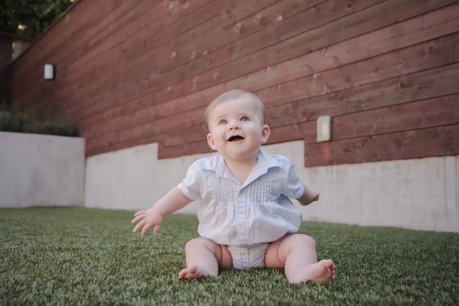Baby boy in a pale blue romper sitting on the grass in a San Francisco backyard, looking up and smiling