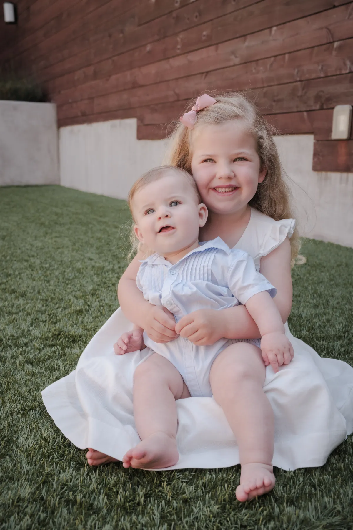 Big sister in a white dress hugging her baby brother on the backyard lawn