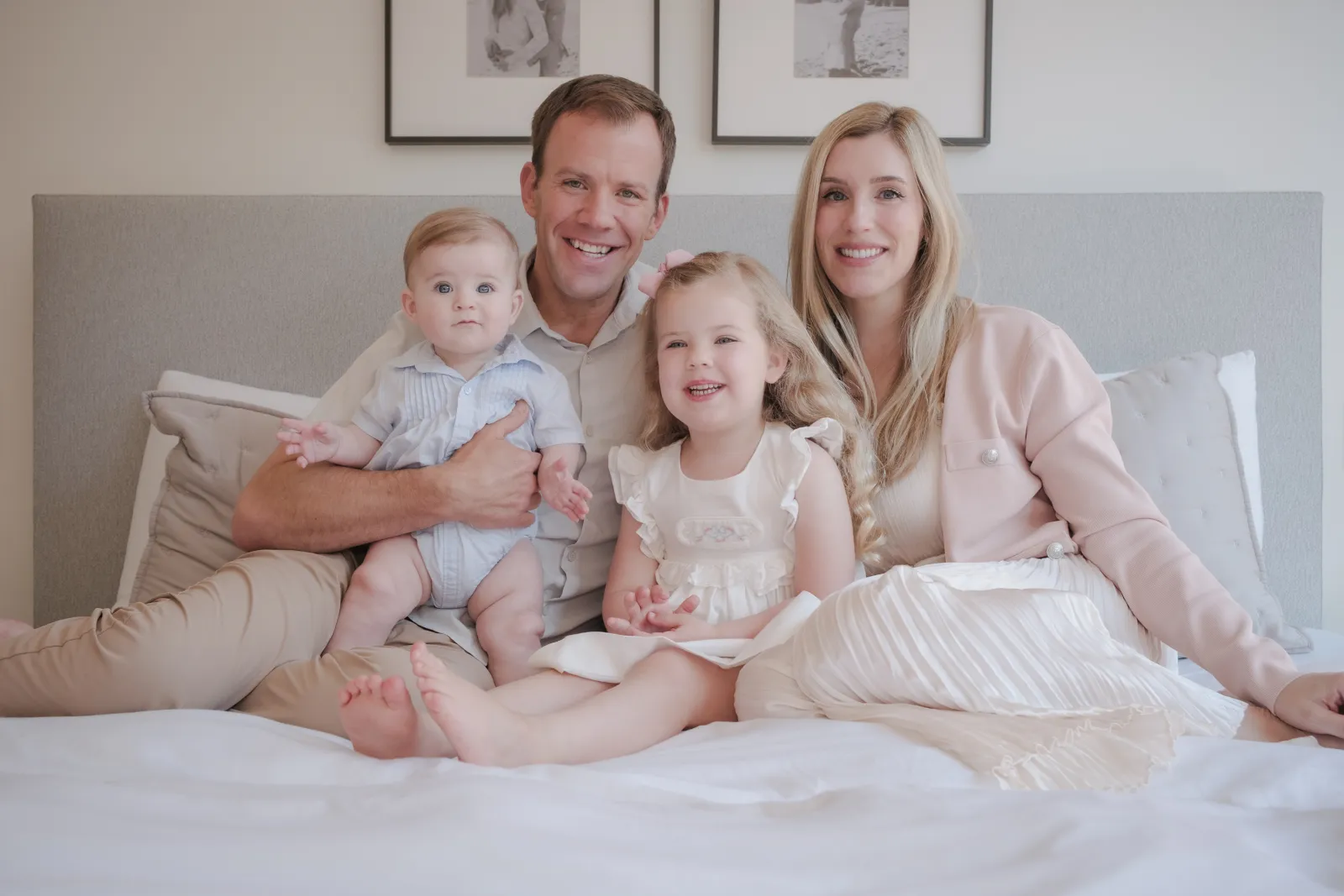 Mom, Dad, daughter and baby son sitting together on a white bed in soft window light