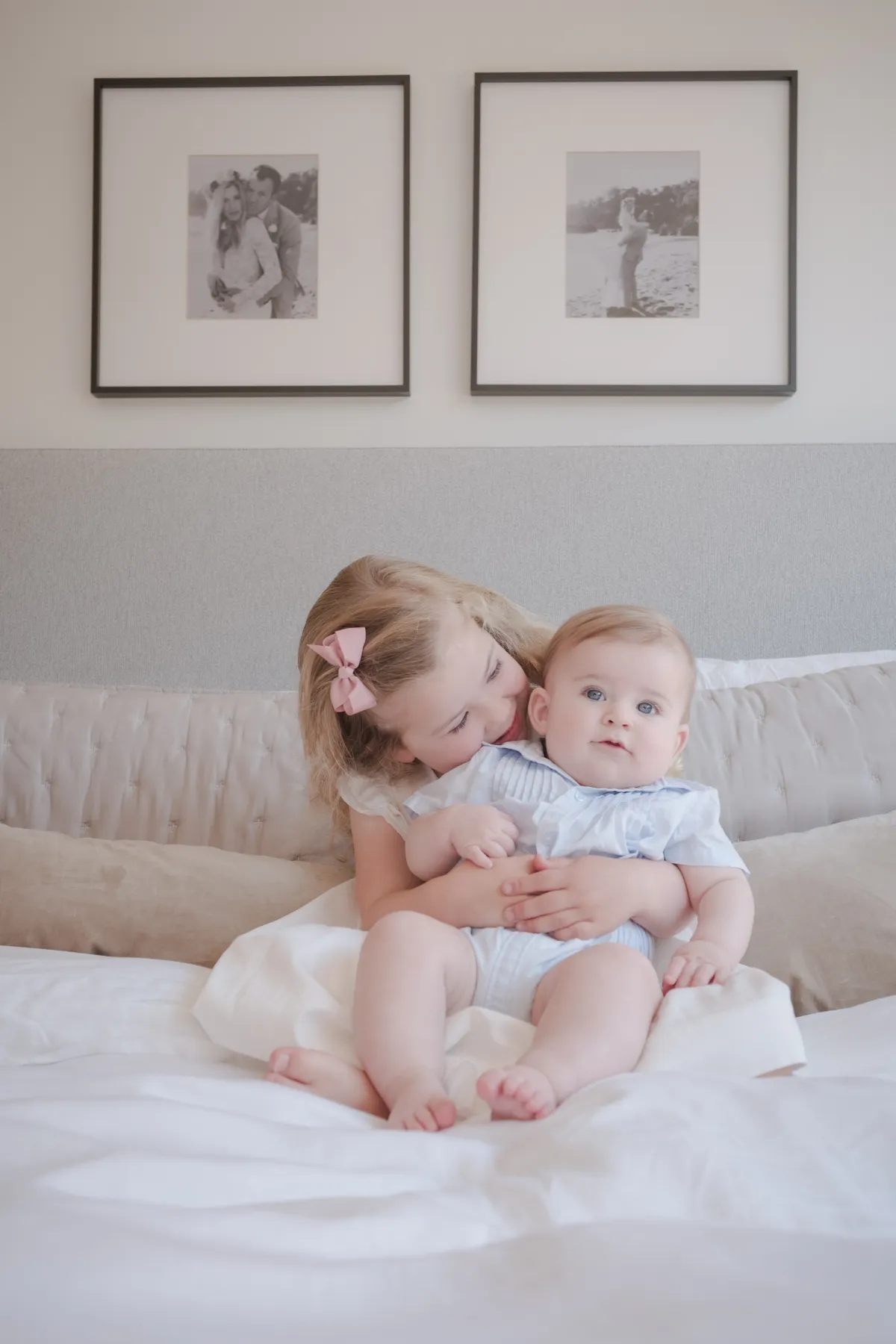 Big sister resting her cheek on her baby brother's head beneath framed black-and-white wedding photos