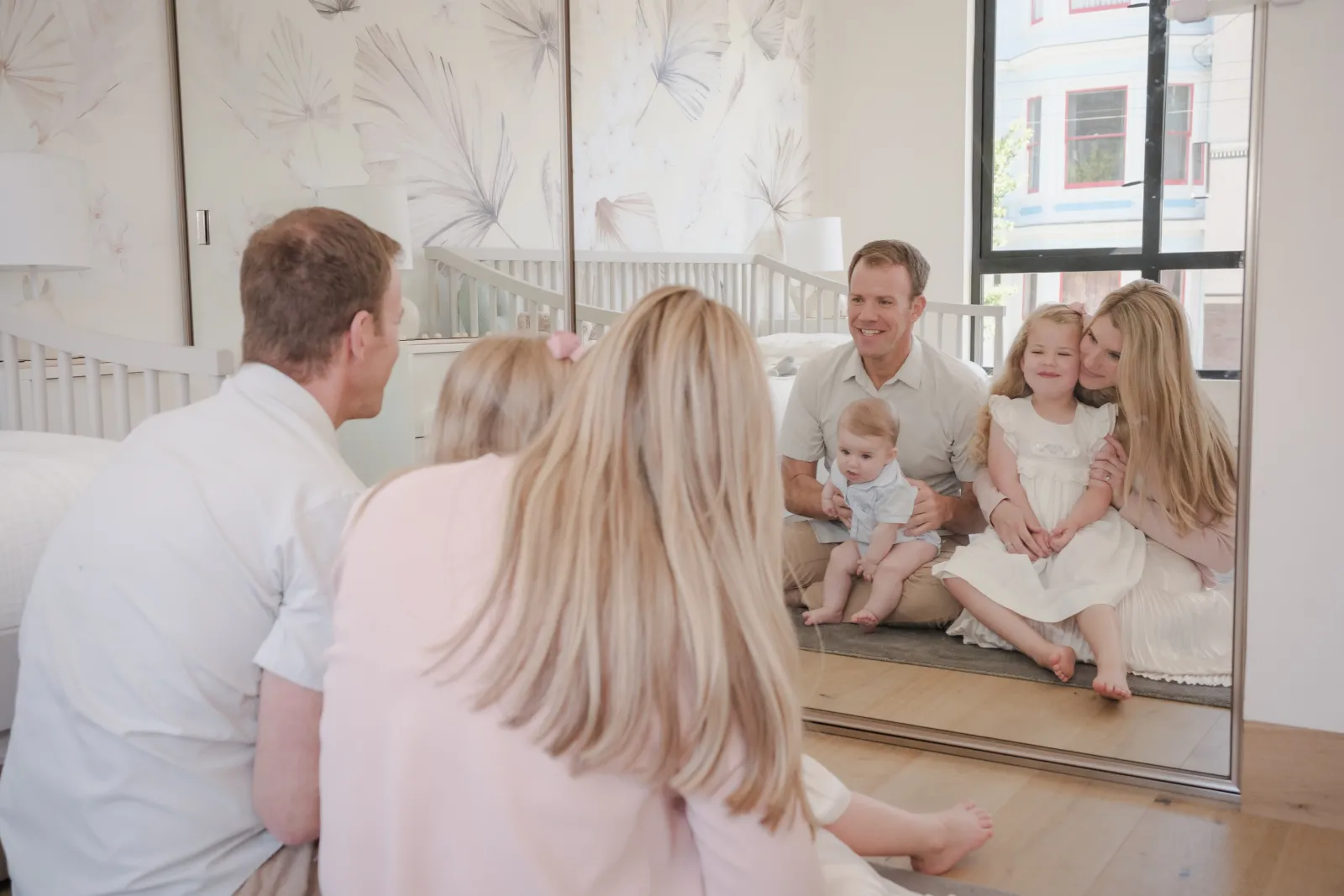 Family reflected in a tall nursery mirror, with Victorian homes visible through the window behind them in Alamo Square