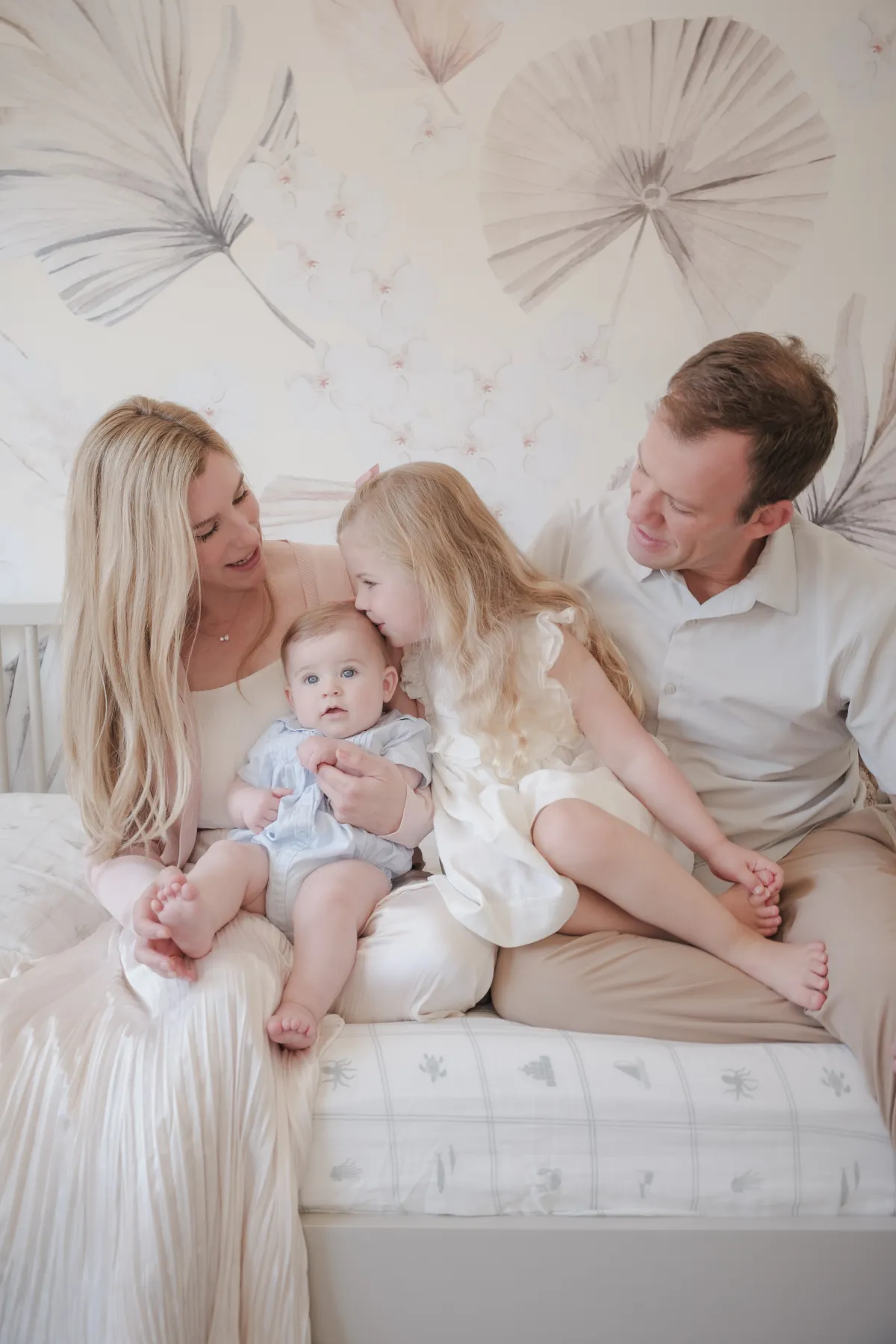 Girl kissing her baby brother's head as her parents look on in a nursery with palm-leaf wallpaper
