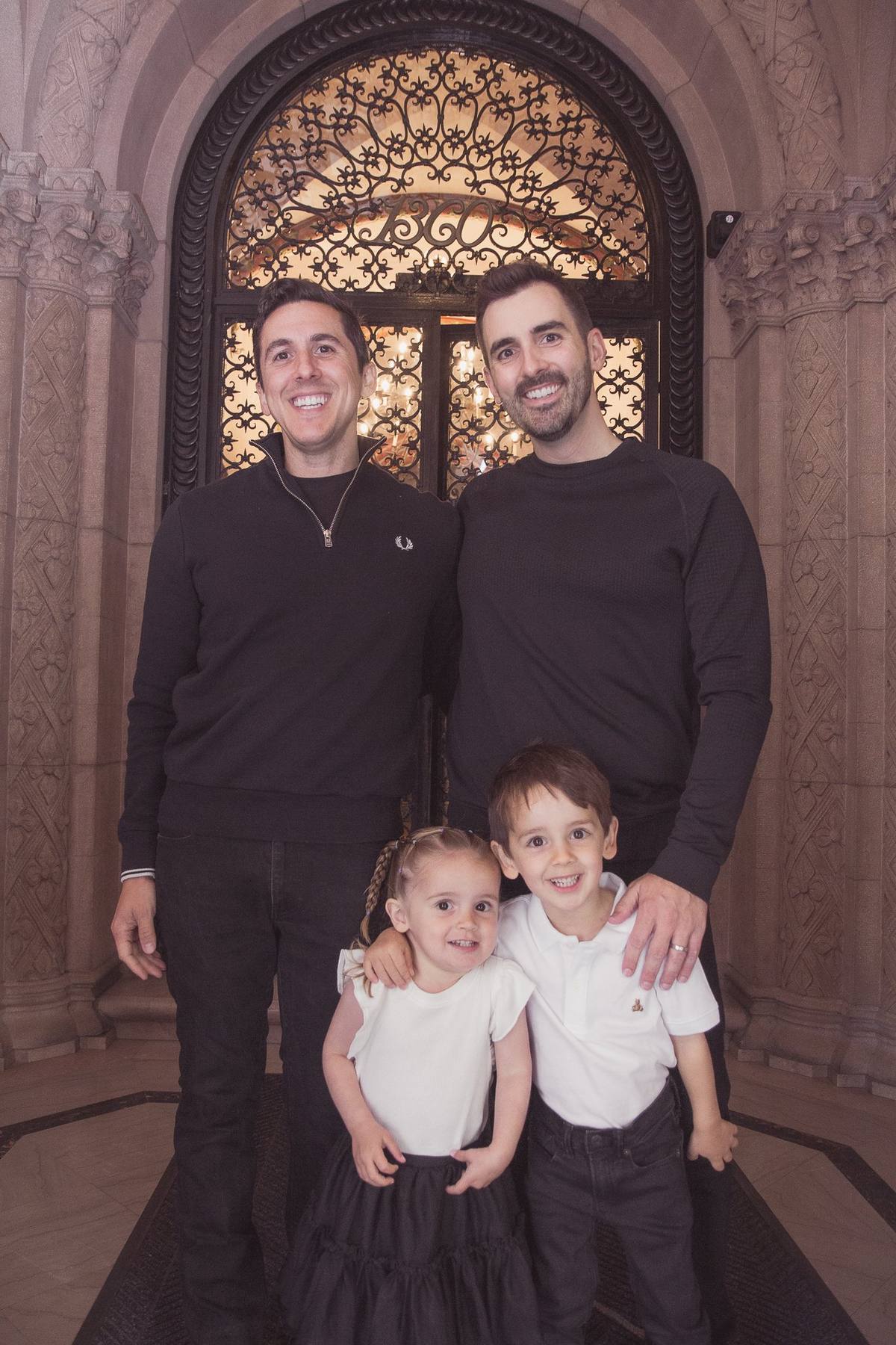 Two dads with their son and daughter in front of the building's arched iron doorway