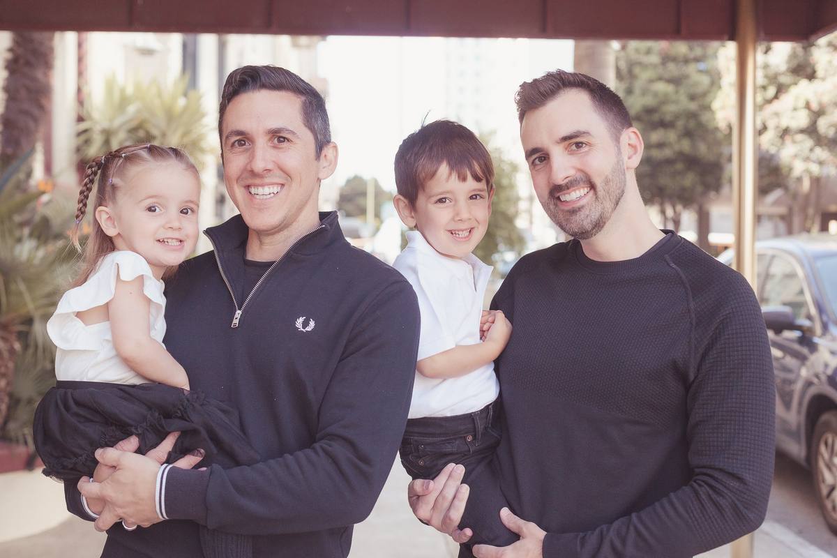 Two dads smiling with their son and daughter in their arms outside their building