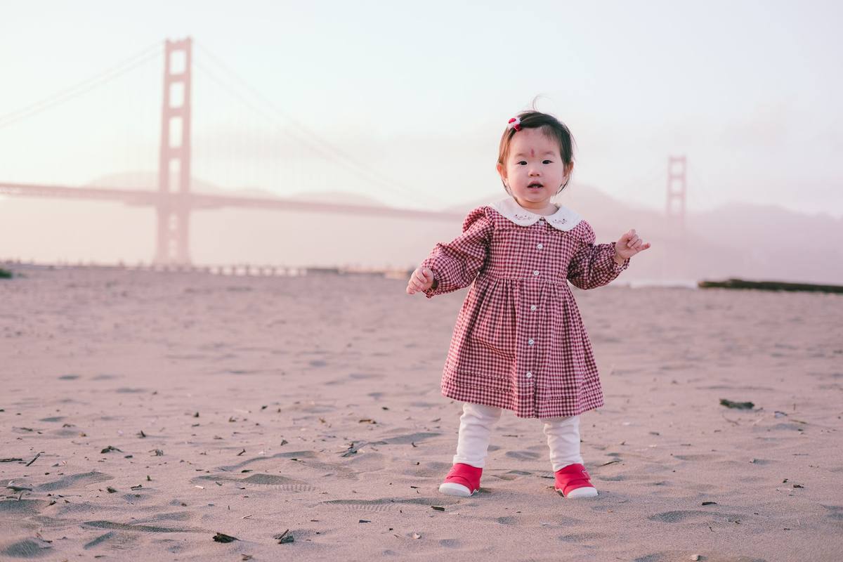 Baby girl in a gingham dress standing on the sand at Crissy Field with the Golden Gate Bridge behind her