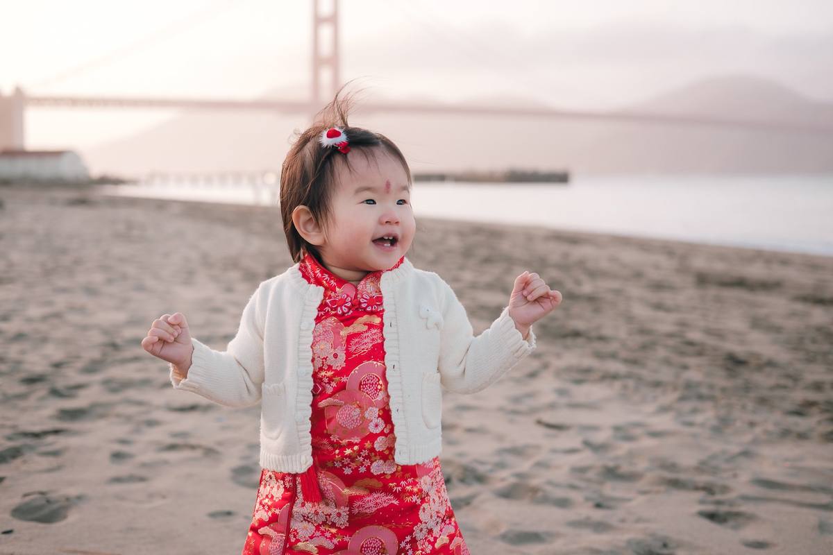 Baby girl in a red brocade dress and cream cardigan laughing on the beach at Crissy Field