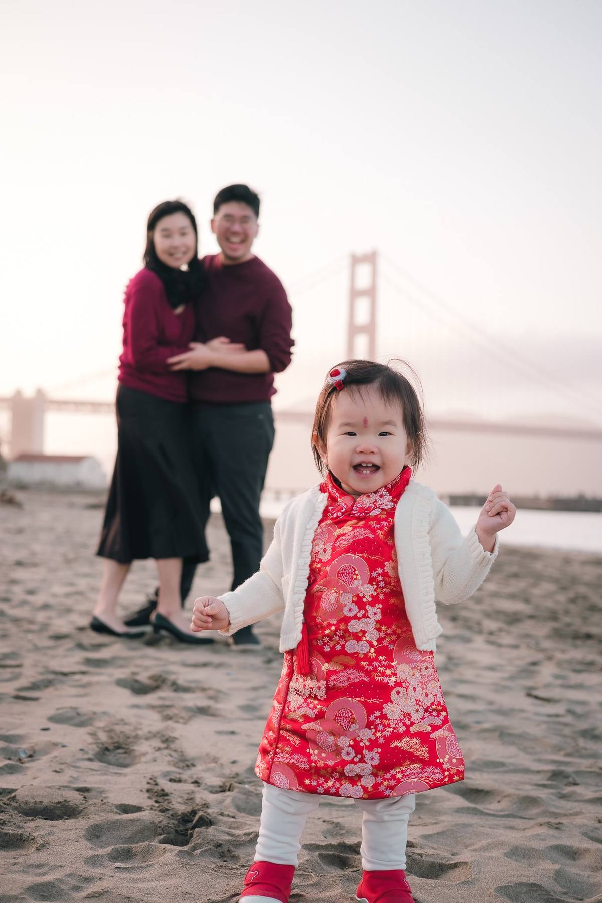 Baby girl in a red brocade dress on the sand with her parents behind her and the Golden Gate Bridge in the distance
