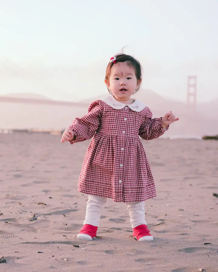 Baby girl in a gingham dress standing on the sand at Crissy Field with the Golden Gate Bridge behind her