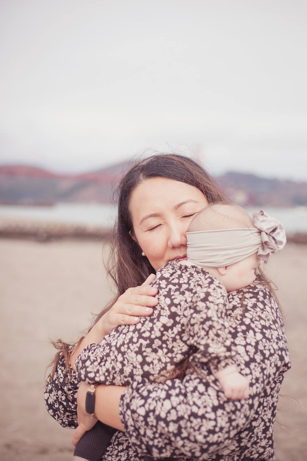 Mother cuddling her baby daughter in a big headband bow on the beach at Crissy Field