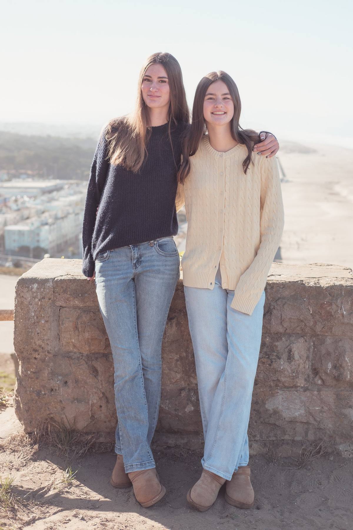 Two teenage sisters leaning on a stone wall with Ocean Beach behind them