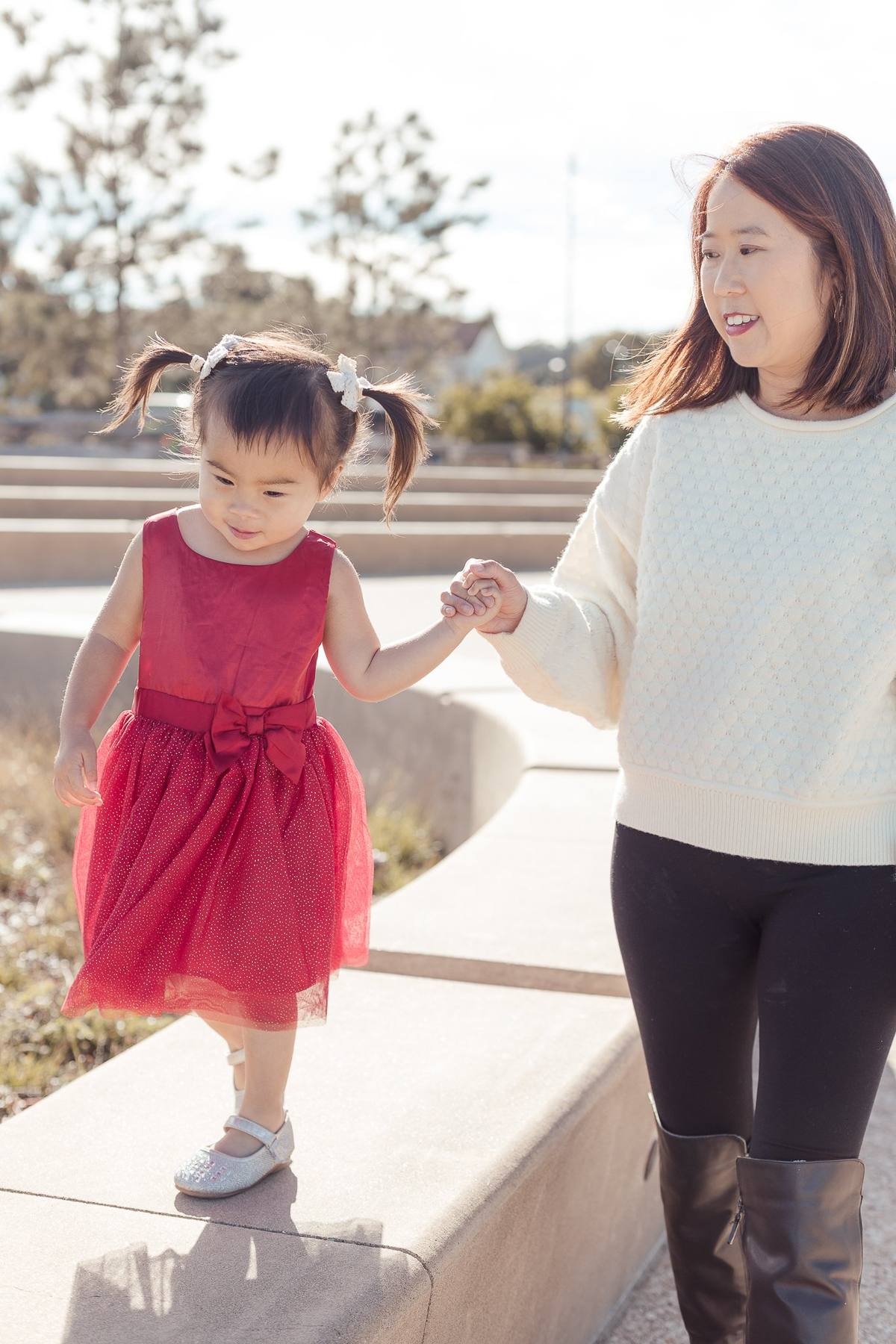 Toddler in a red dress holding her mother's hand as she walks along a low wall