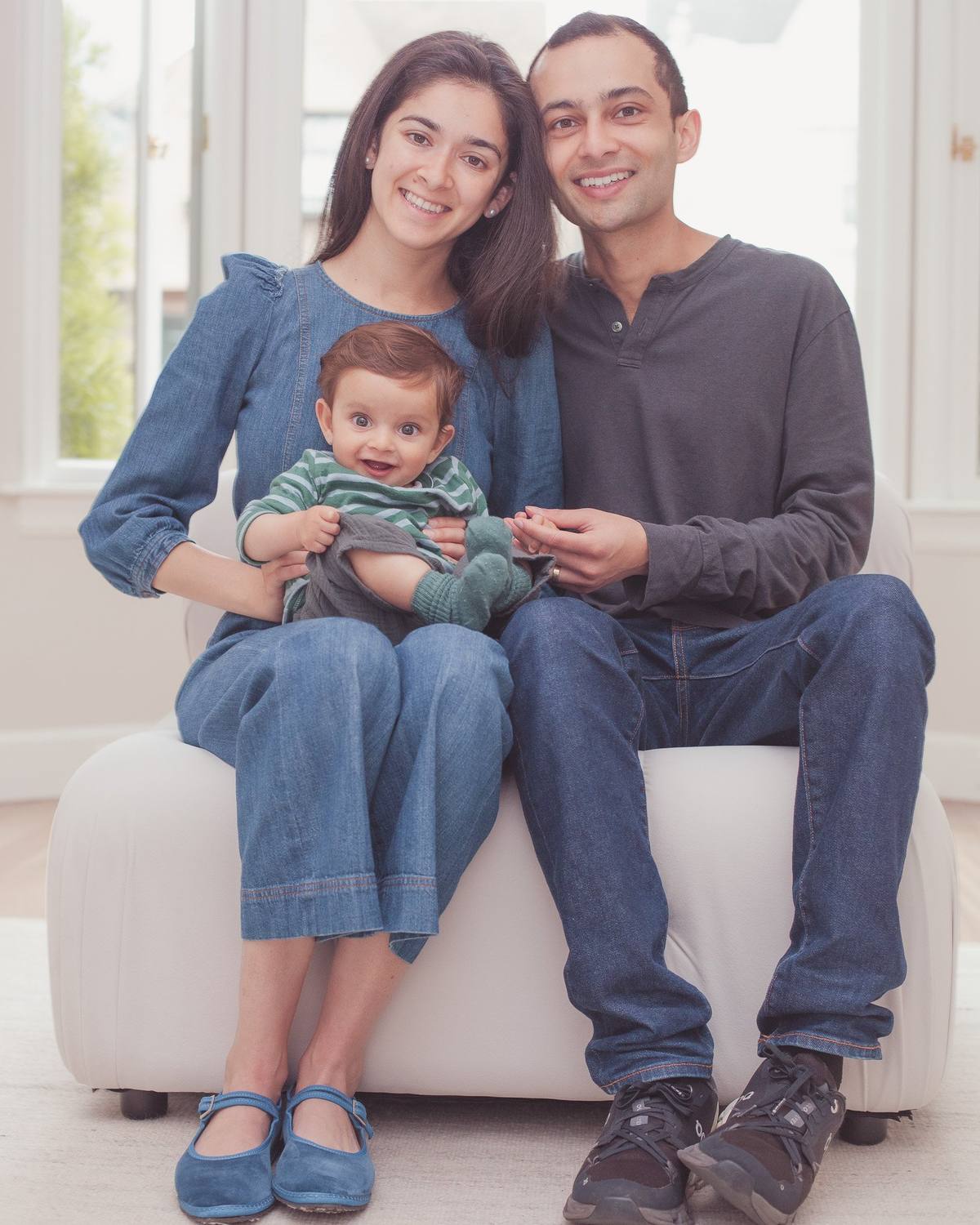 Parents and their baby boy sitting together on a cream armchair at home, dressed in soft denim and gray