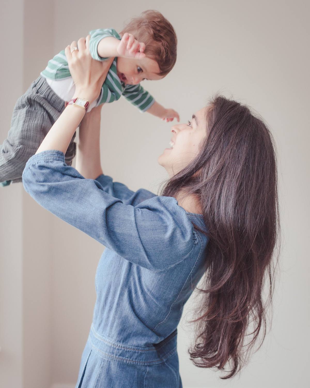 Mother lifting her baby son overhead in the soft window light of their home