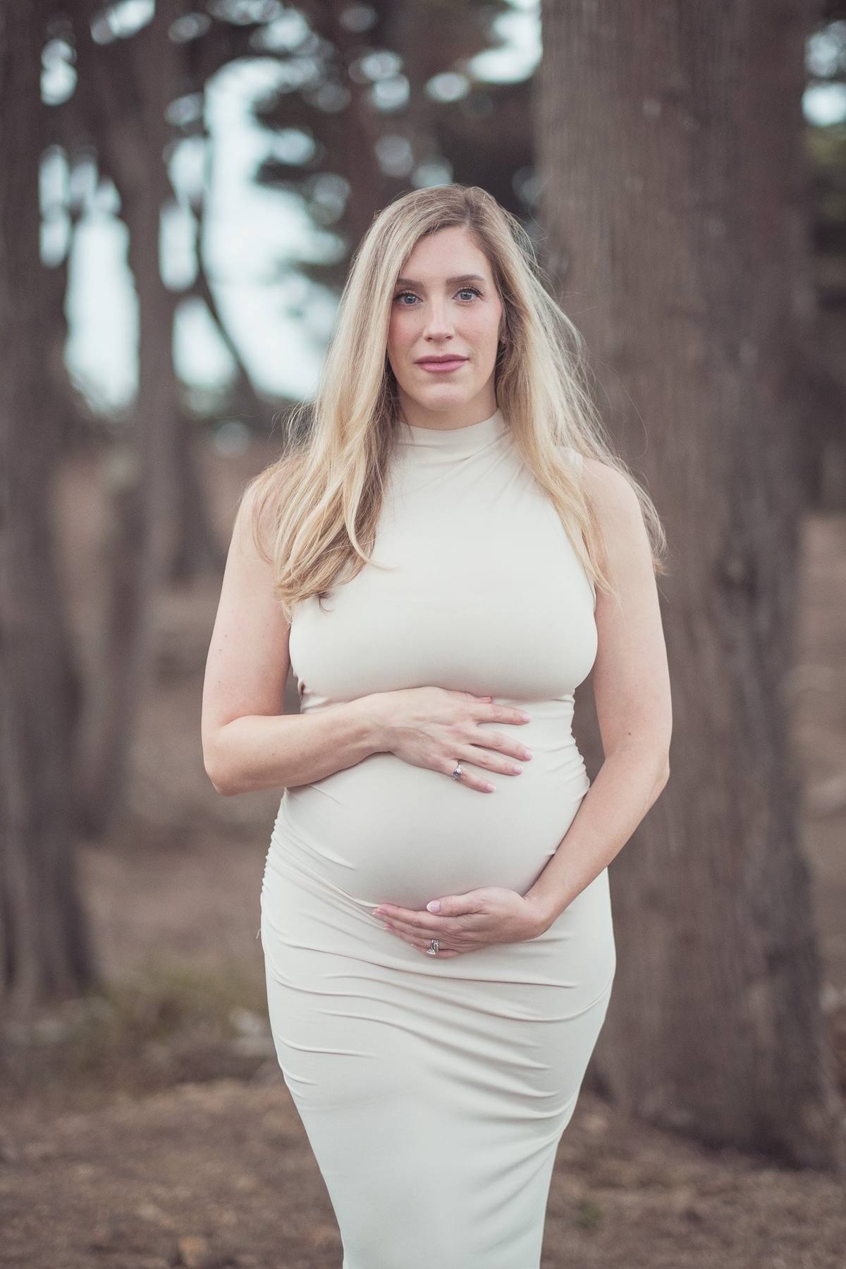 Pregnant mother in a cream dress standing among the tall trees at Lands End
