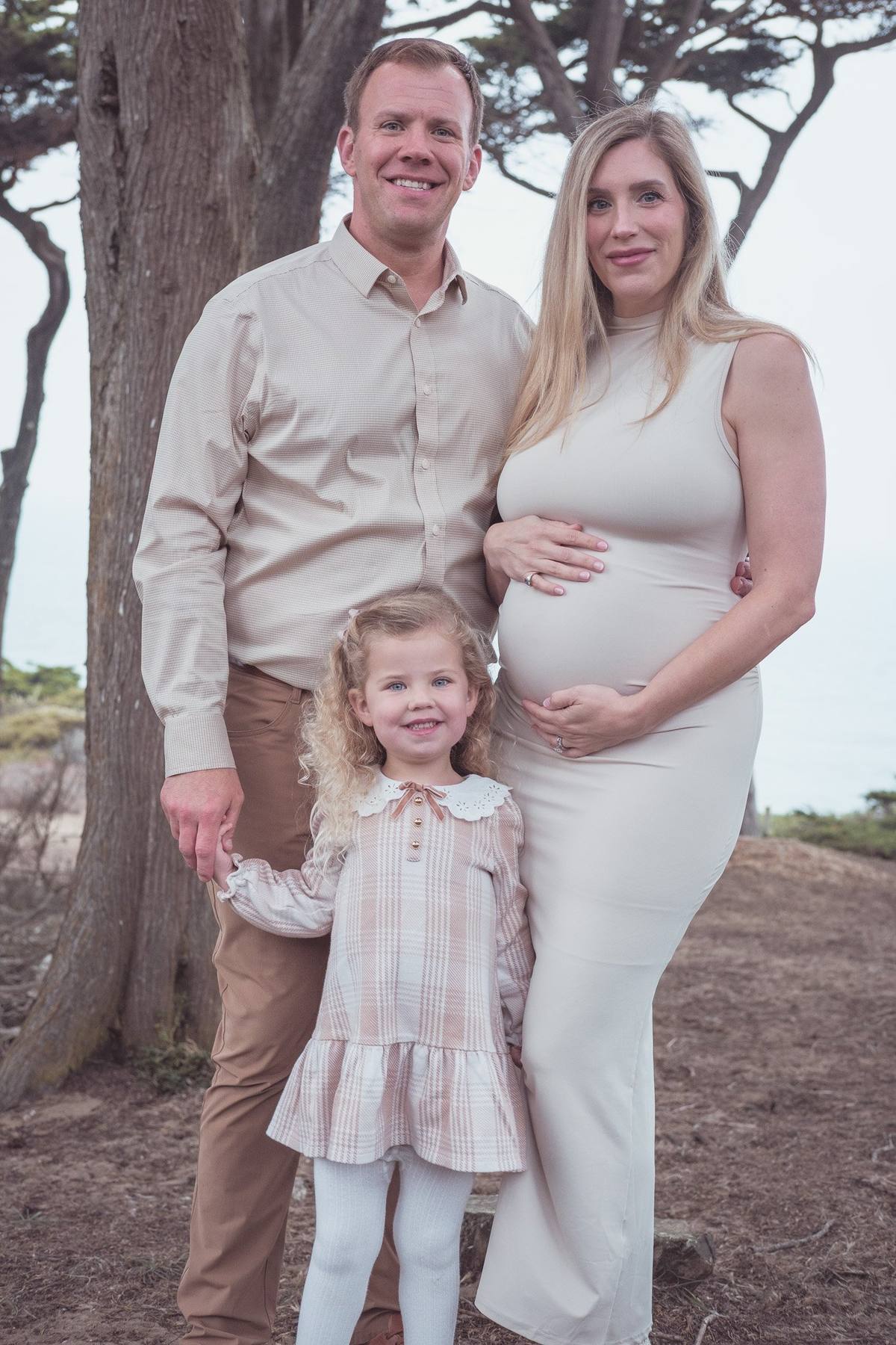 Expecting parents and their young daughter standing together among the trees at Lands End