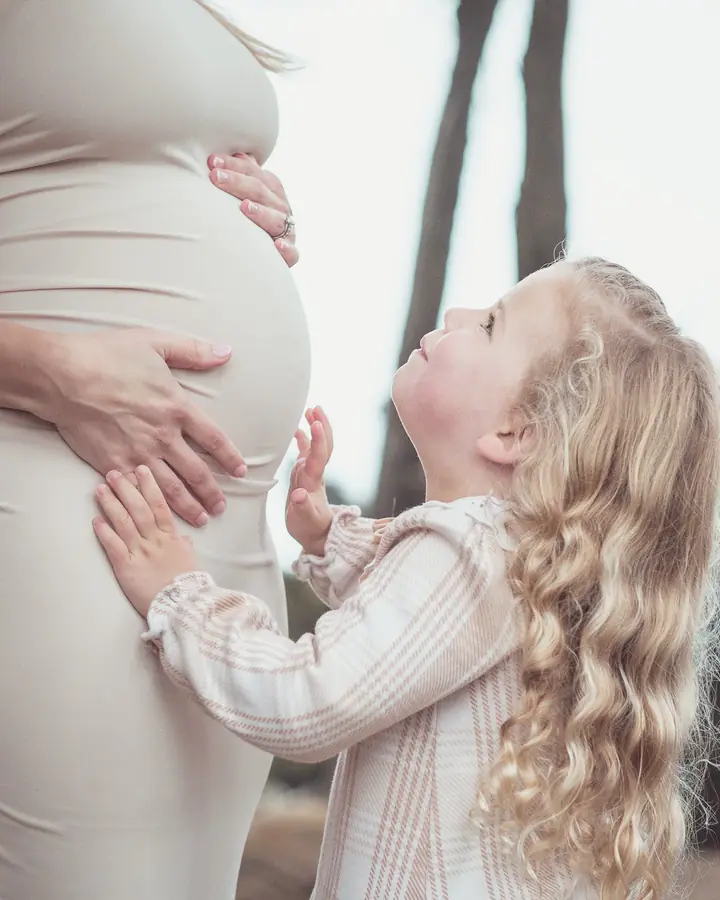 Young daughter reaching up to kiss her pregnant mother's belly at Lands End
