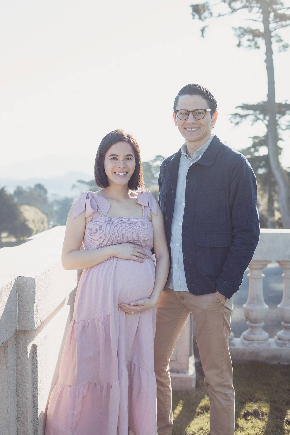 Pregnant woman in a pink dress smiling beside her husband at the Legion of Honor balustrade