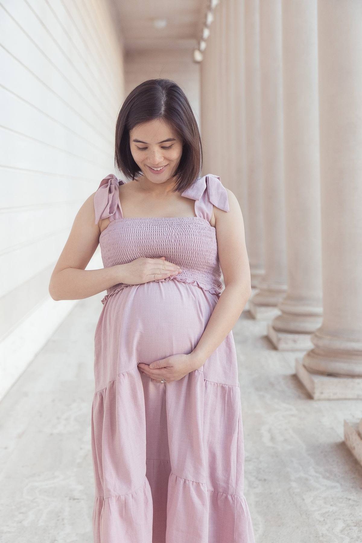 Pregnant woman cradling her bump in a pink tiered dress between the columns
