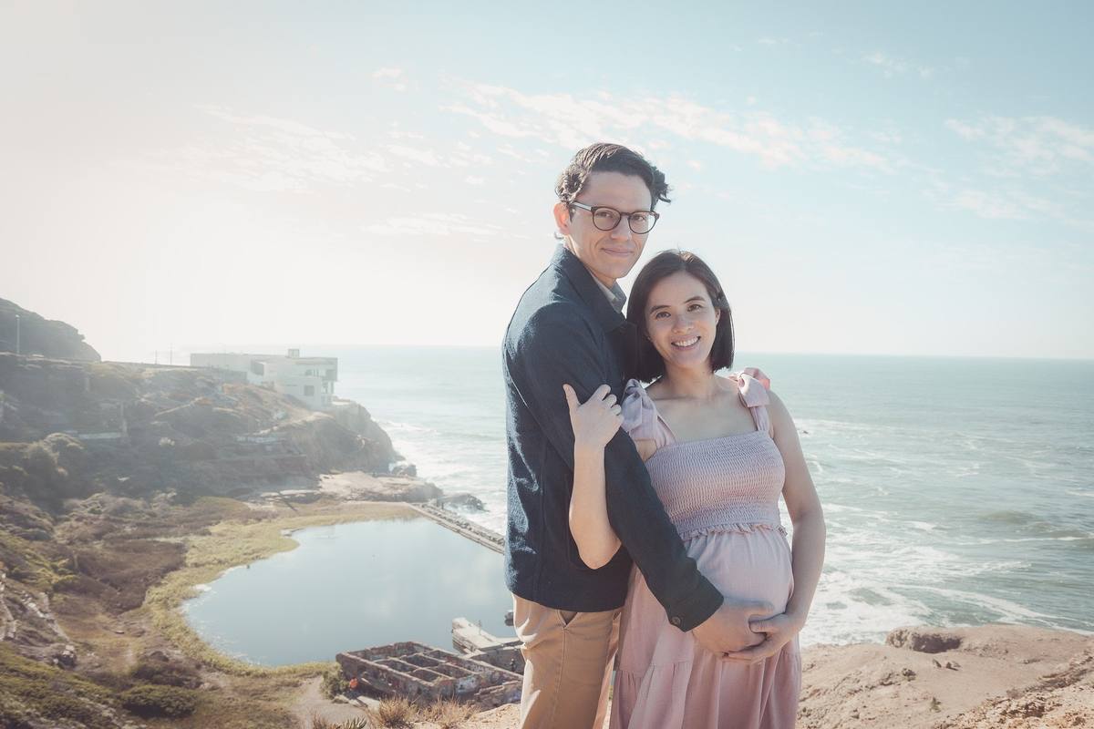 Expecting couple smiling on the bluff at Lands End with the Sutro Baths and the Pacific behind them