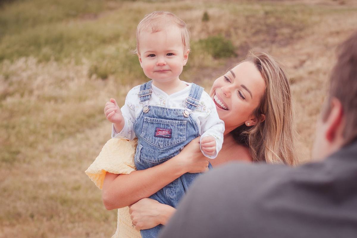 Baby boy in denim overalls smiling in his mother's arms on a grassy hillside