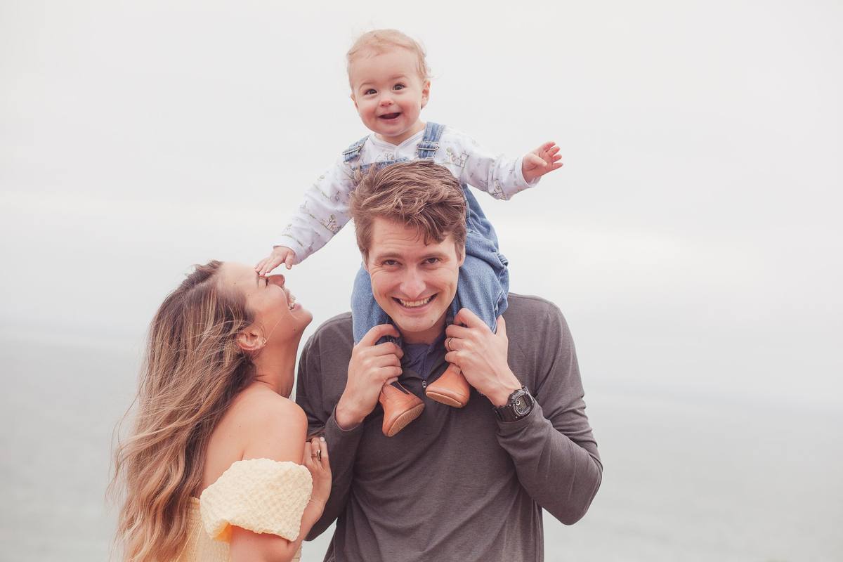 Baby boy on his father's shoulders reaching for mom with a foggy ocean horizon behind them