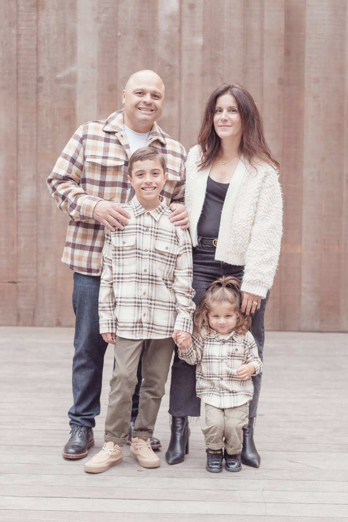 Family of four in coordinated plaid, cream, and denim outfits standing in front of a weathered wood wall