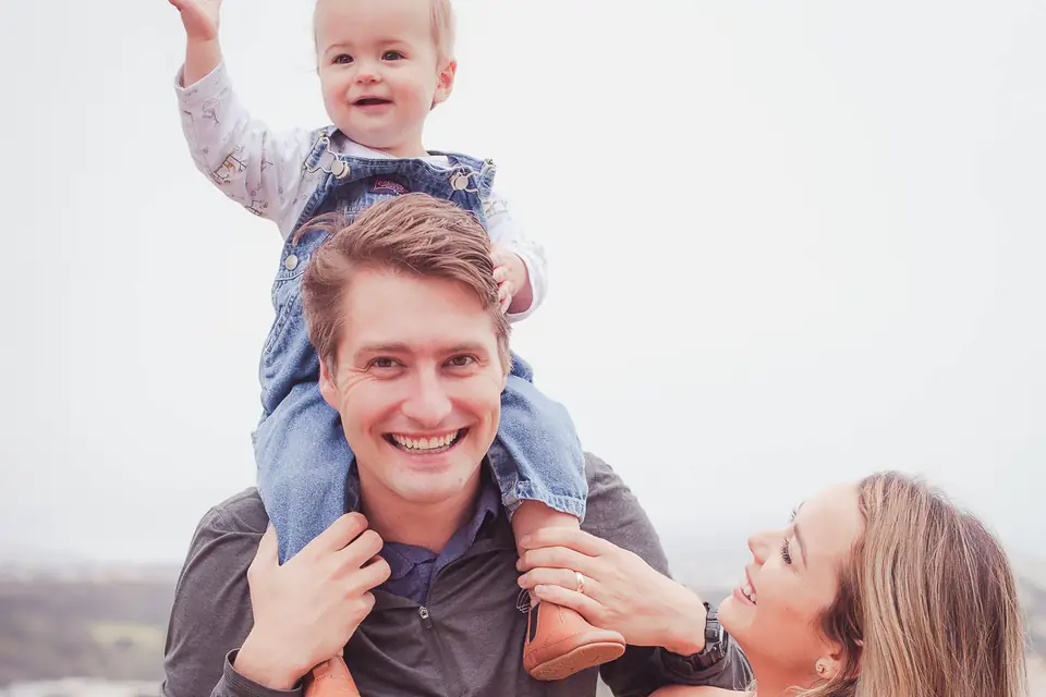 Dad in a charcoal pullover carrying his son on his shoulders beside mom in a pale yellow dress