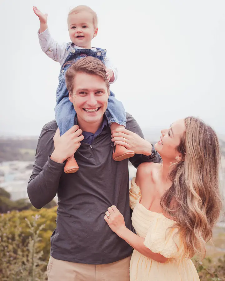 Dad in a charcoal pullover carrying his son on his shoulders beside mom in a pale yellow dress