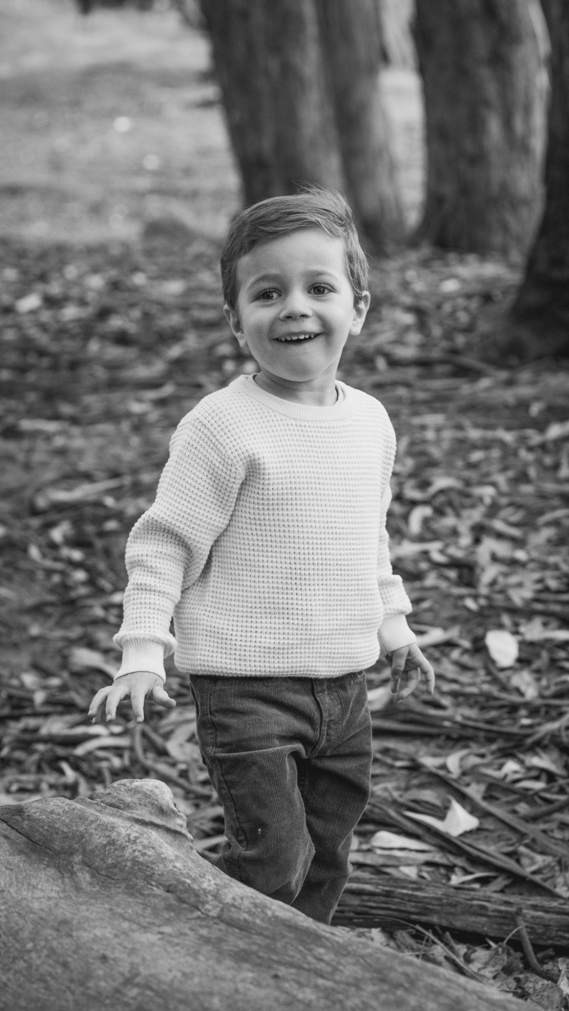 Little boy in a knit sweater grinning among the eucalyptus at Andy Goldsworthy&rsquo;s Wood Line in the Presidio, black and white