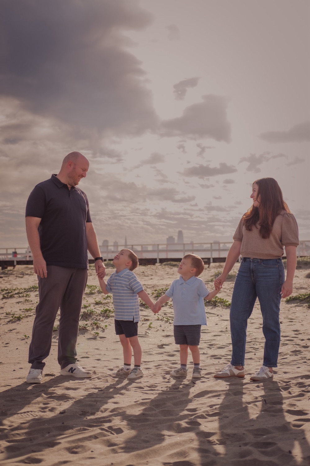 Family of four holding hands on the beach at Crissy Field with the San Francisco skyline behind them, family photography by Matt Dea