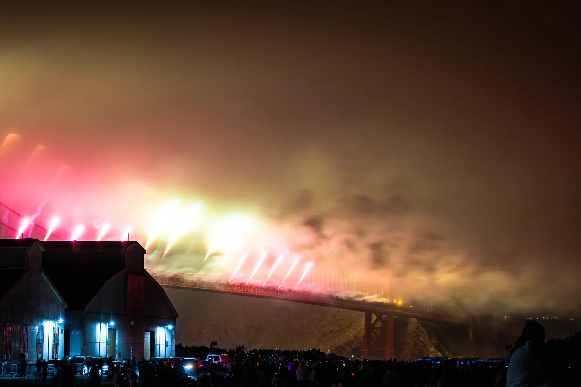 Fireworks pouring off the Golden Gate Bridge into the fog, seen past a Crissy Field hangar and the crowd