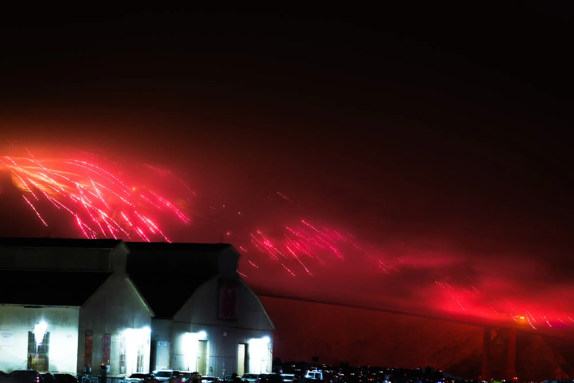 Red fireworks streaking along the fog-hidden bridge above a lit Crissy Field hangar